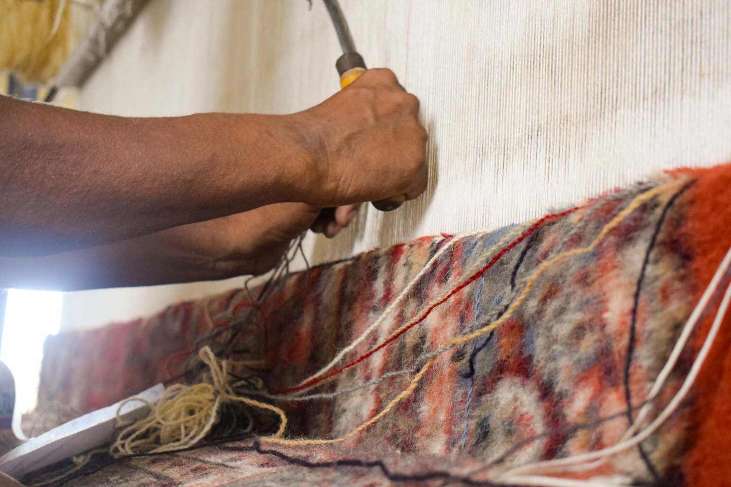 Hands use a tool to weave threads into a rug.