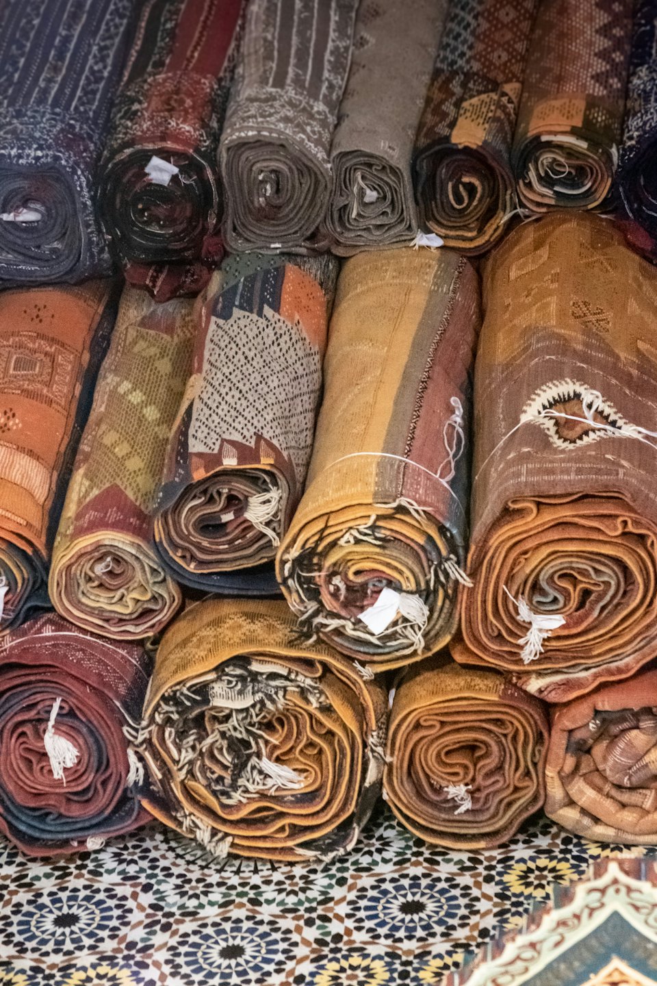 Rolled hand-woven rugs with geometric patterns stacked on a mosaic surface.